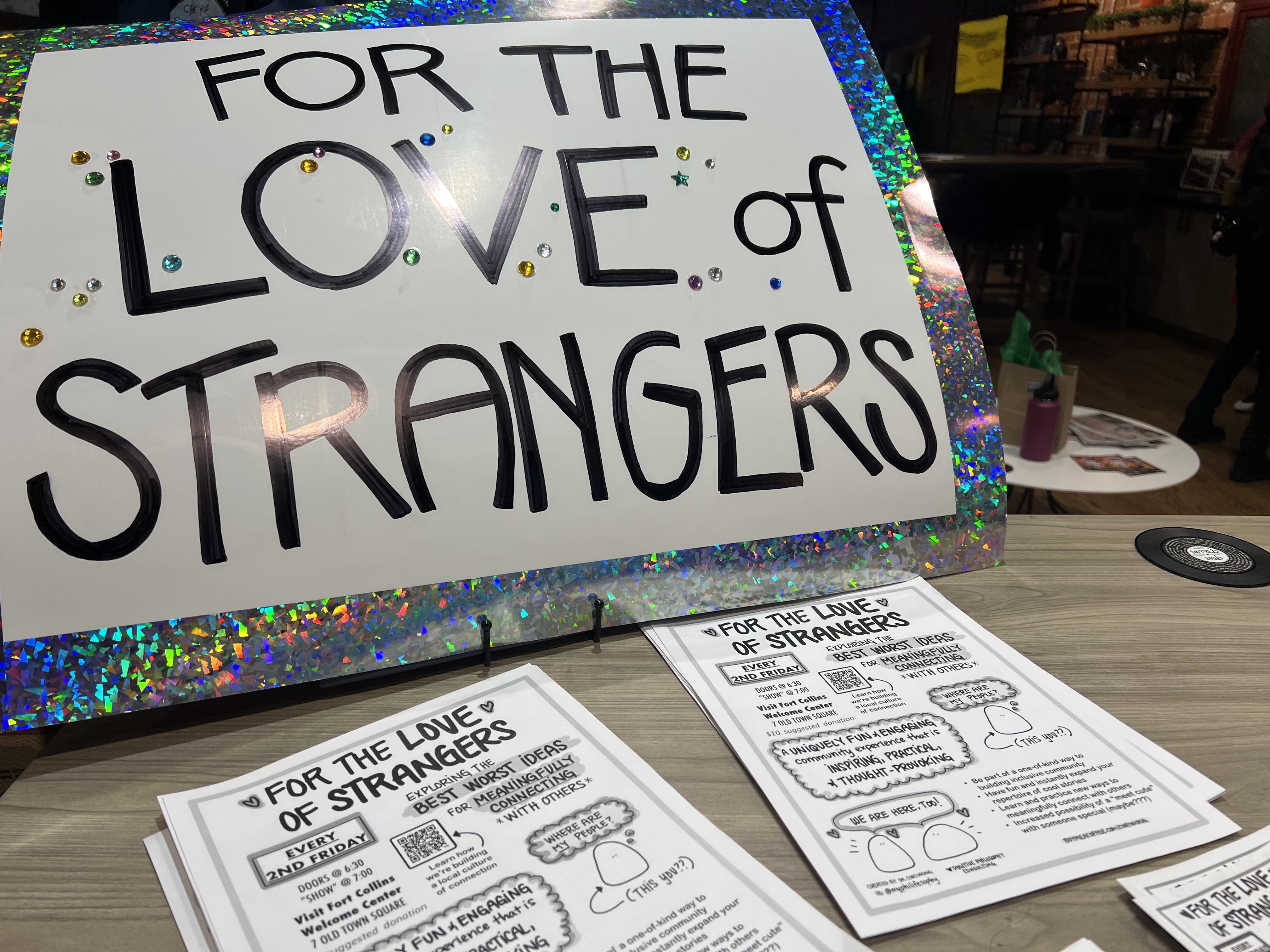 Handmade posterboard sign and paper fliers on a table promoting For The Love of Strangers by Dr. Cori Wong