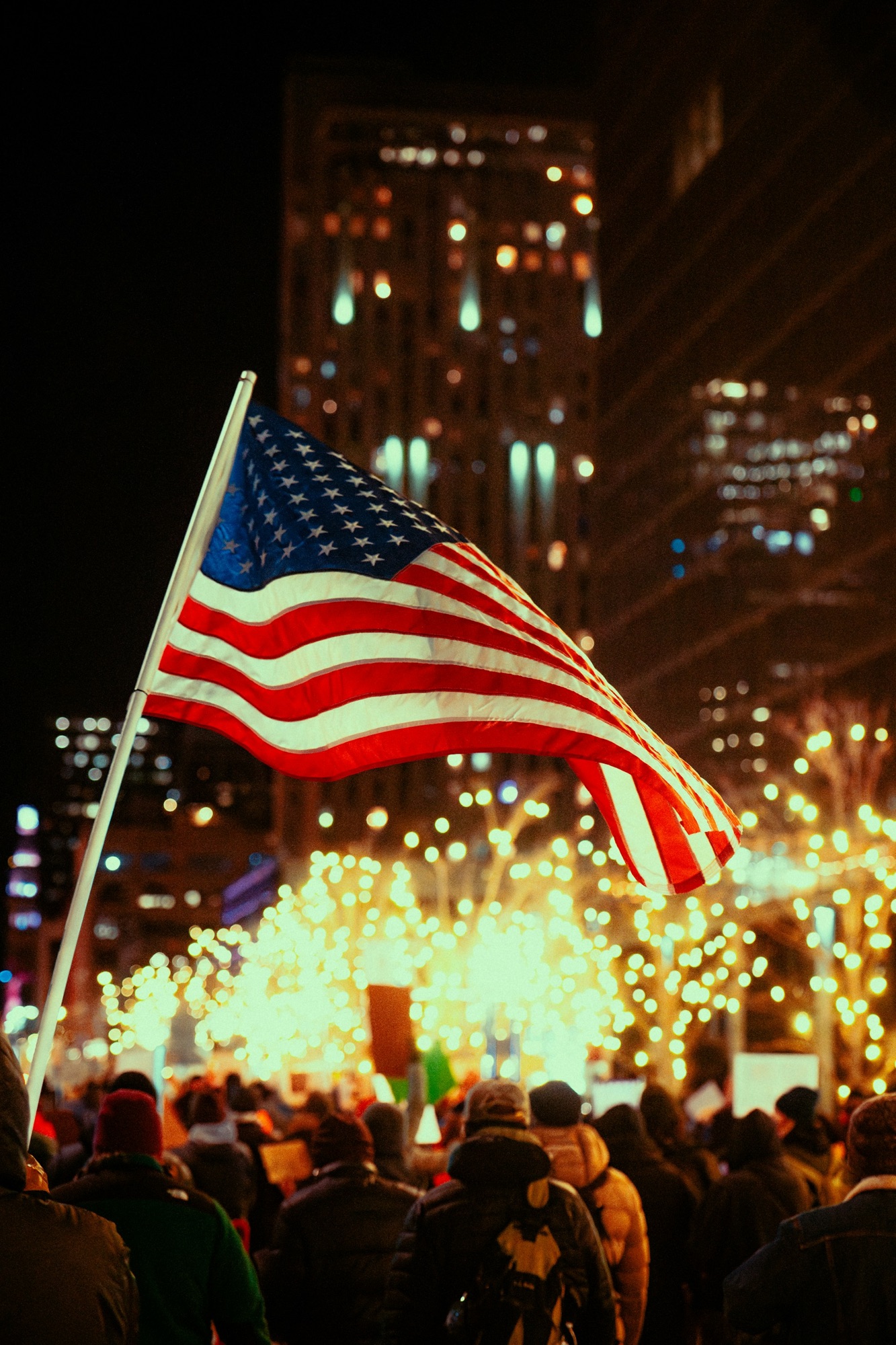 American flag in front of white holiday lights against buildings at night in Denver. Photo by Colin Lloyd/Unsplash
